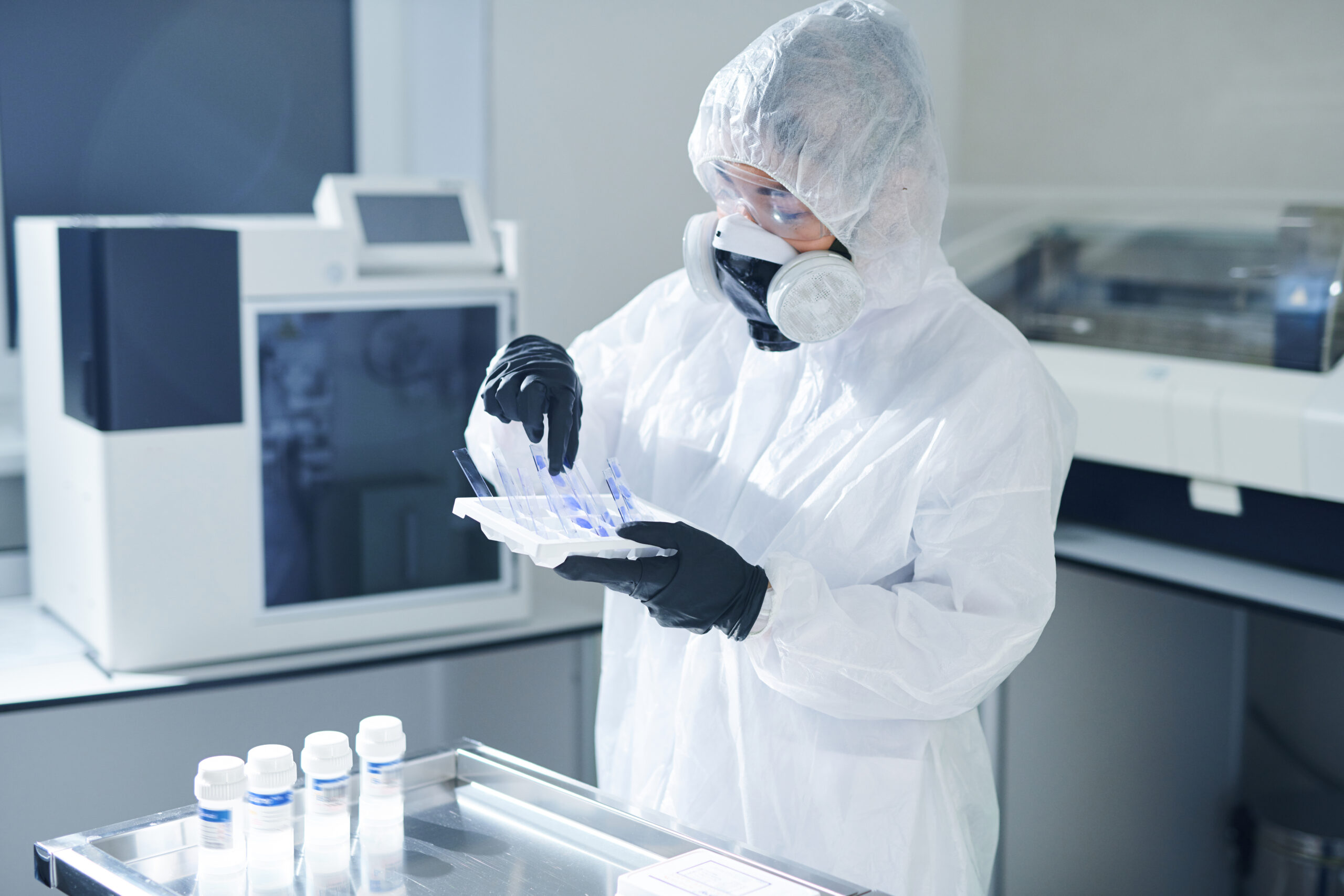 Busy infectious disease scientist in biohazard suit and respiratory mask standing at table with bottles and taking samples of biohazards for research