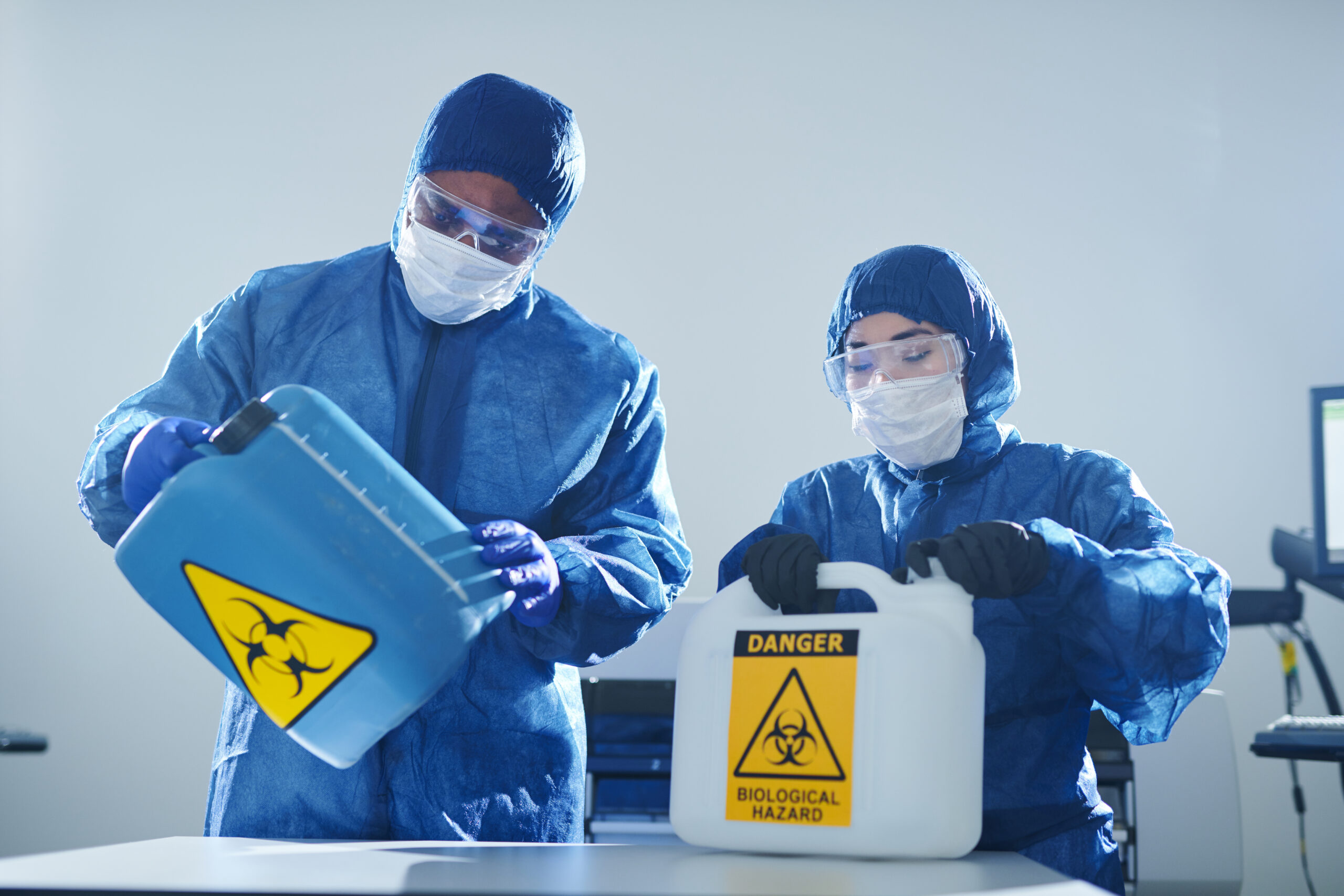 Concentrated multi-ethnic toxic laboratory colleagues in protective suits and masks standing at table and opening gallons with biohazard symbols while working with biohazards